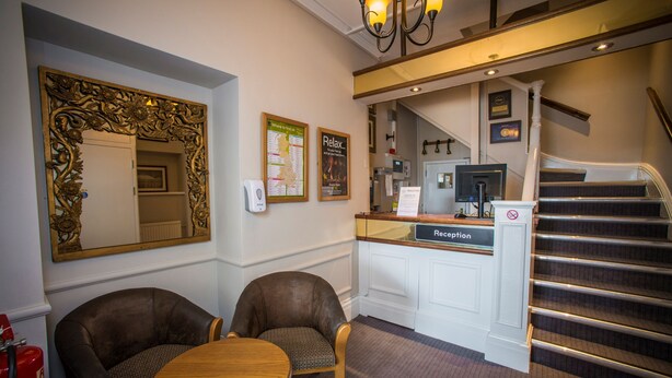 A reception area with a reception desk, two armchairs, a round table, a large ornate mirror, and a staircase. - Old Colonial Hotel Weston-Super-Mare | Marston's Inns (Weston-super-Mare)