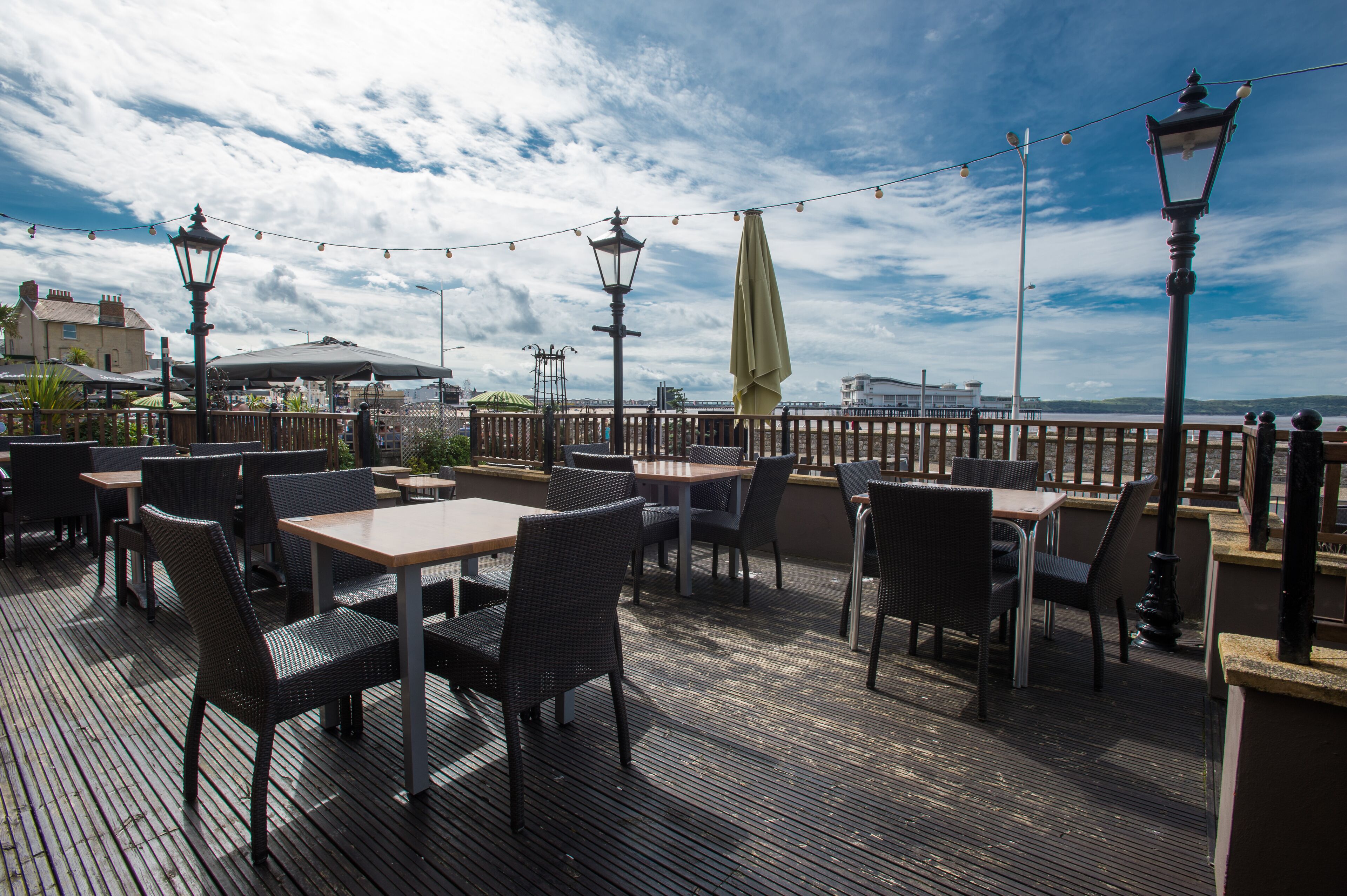 An outdoor dining area with tables and chairs, string lights, and a view of the sea.