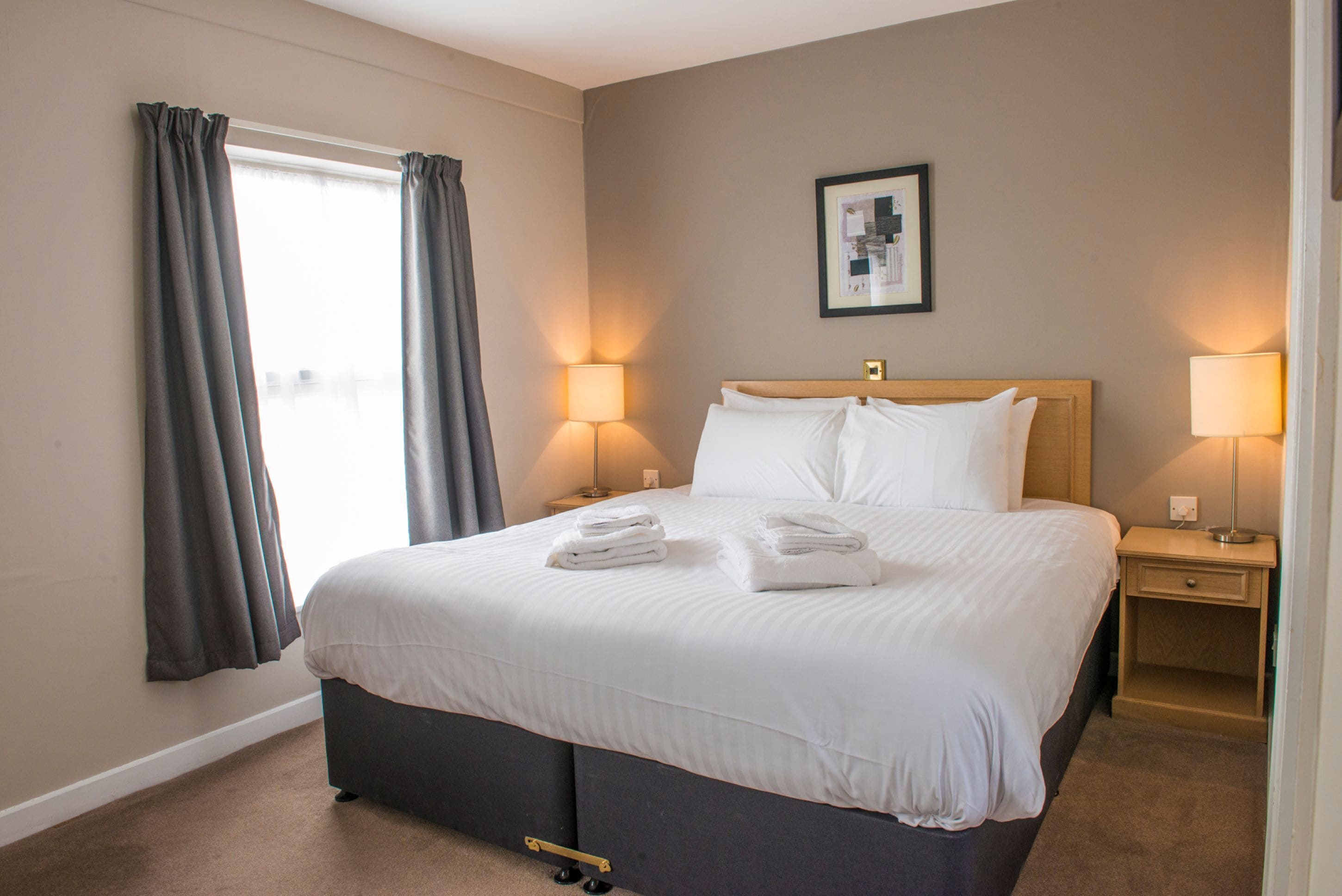 A hotel room with a large bed, two bedside tables with lamps, a window with curtains, and a framed picture on the wall.