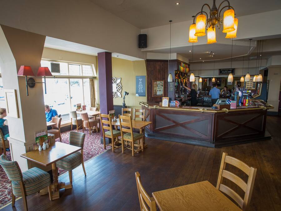 A restaurant interior with wooden tables and chairs, a bar area, and a view of the outdoors through large windows.