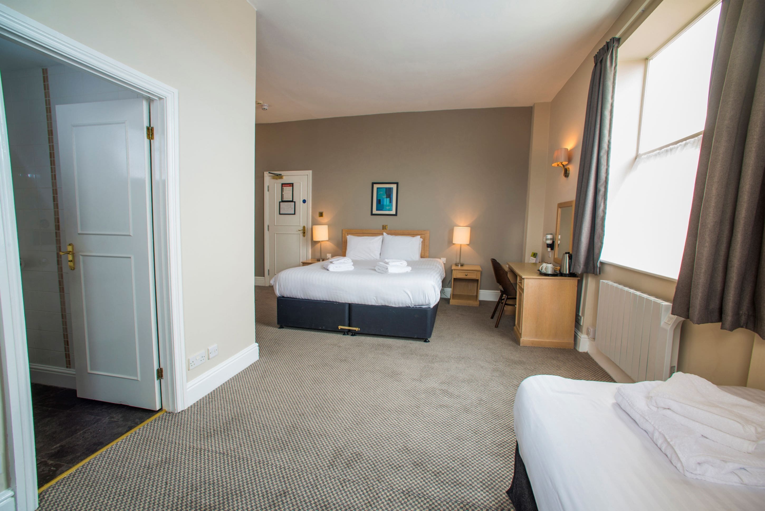 Quarto de hotel com uma cama grande, uma cama menor, uma escrivaninha com cadeira, luminária de mesa, quadro na parede e janela com cortinas.