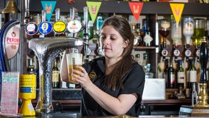 Ein Barkeeper zapft ein Bier an einer Bar mit mehreren Zapfhähnen und verschiedenen Flaschen im Hintergrund.