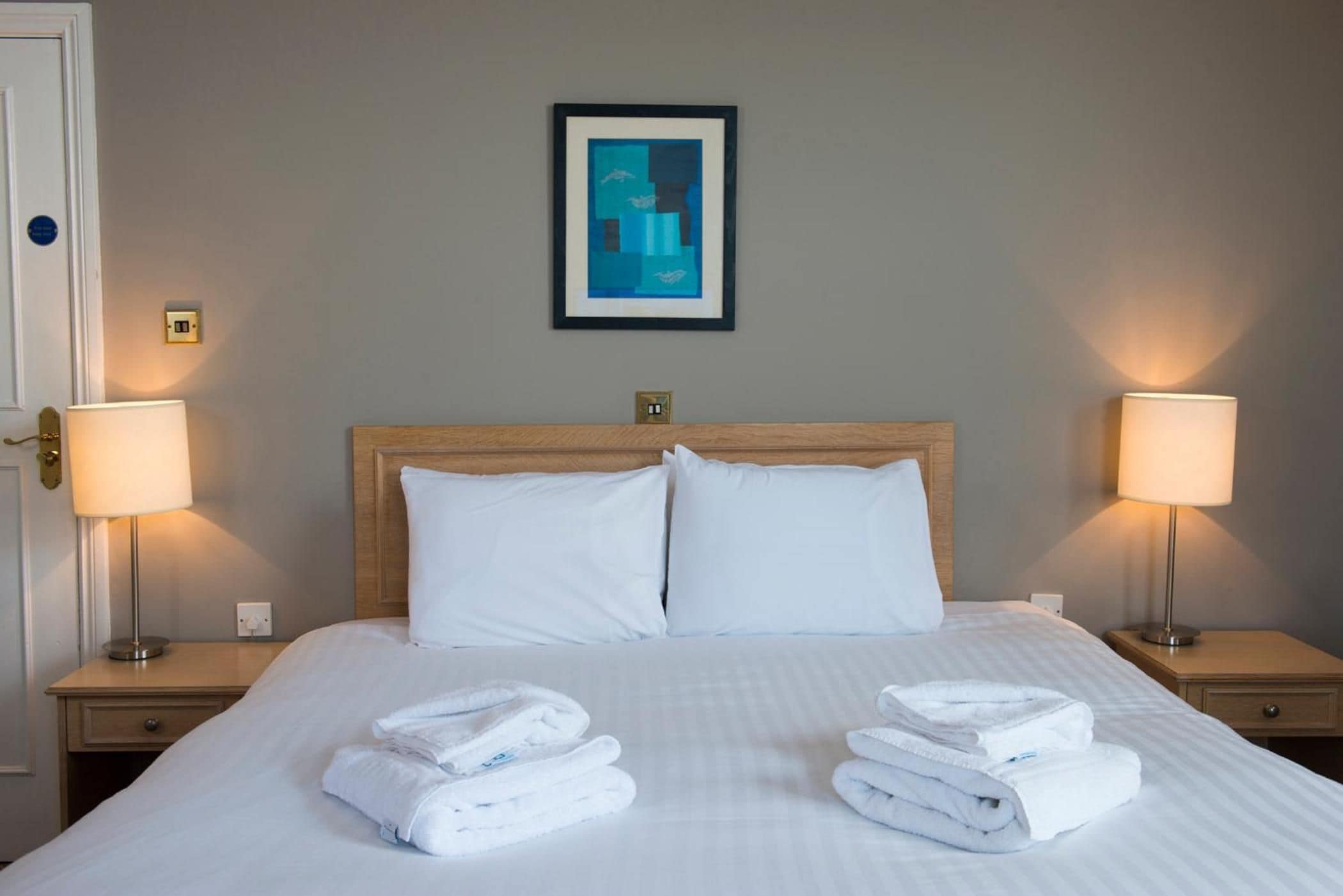 A hotel room with a bed, two bedside lamps, a framed picture on the wall, and folded towels on the bed.
