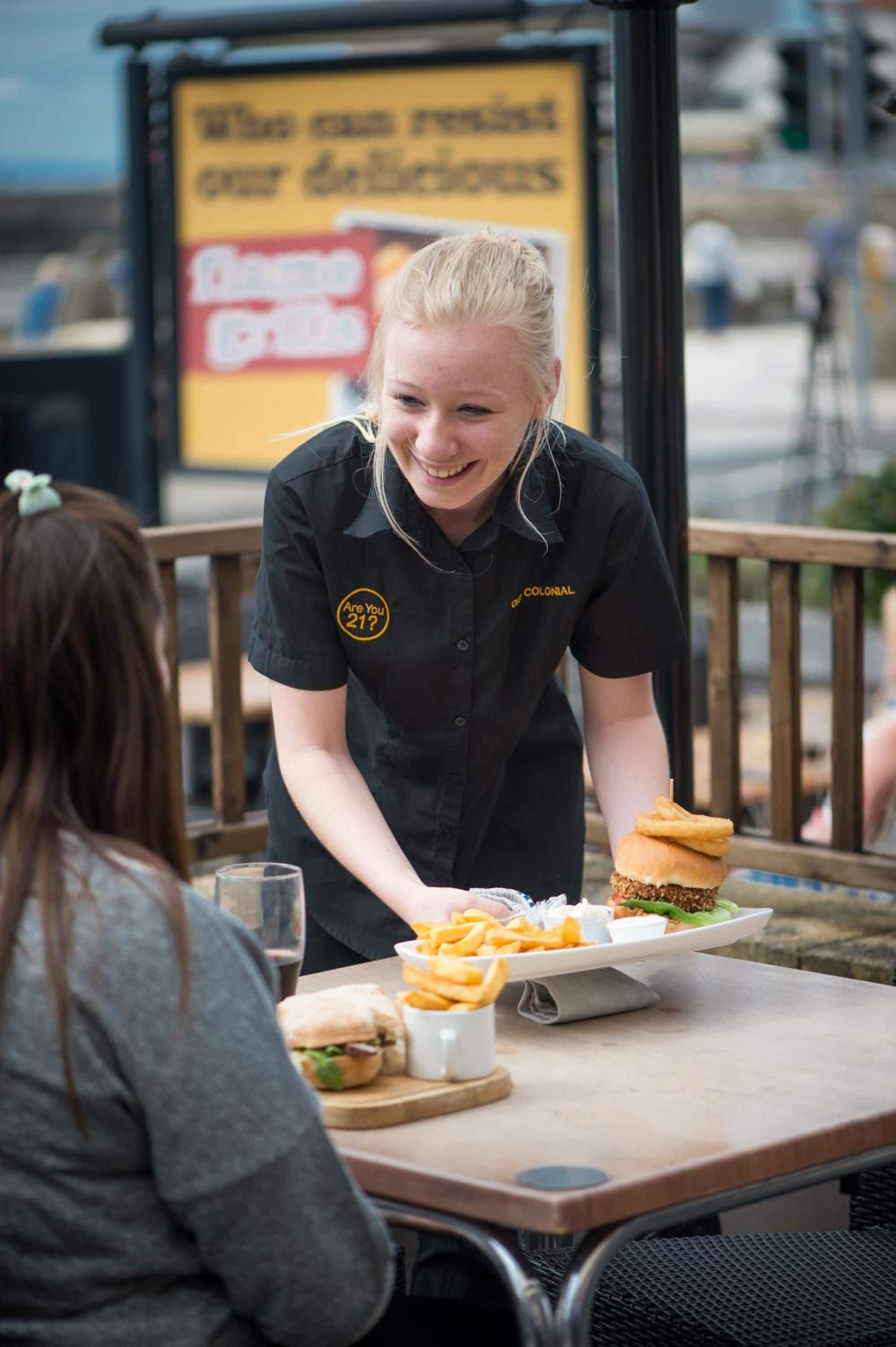 Une personne servant des plats à une table en plein air : on y voit un hamburger, des frites et une boisson.