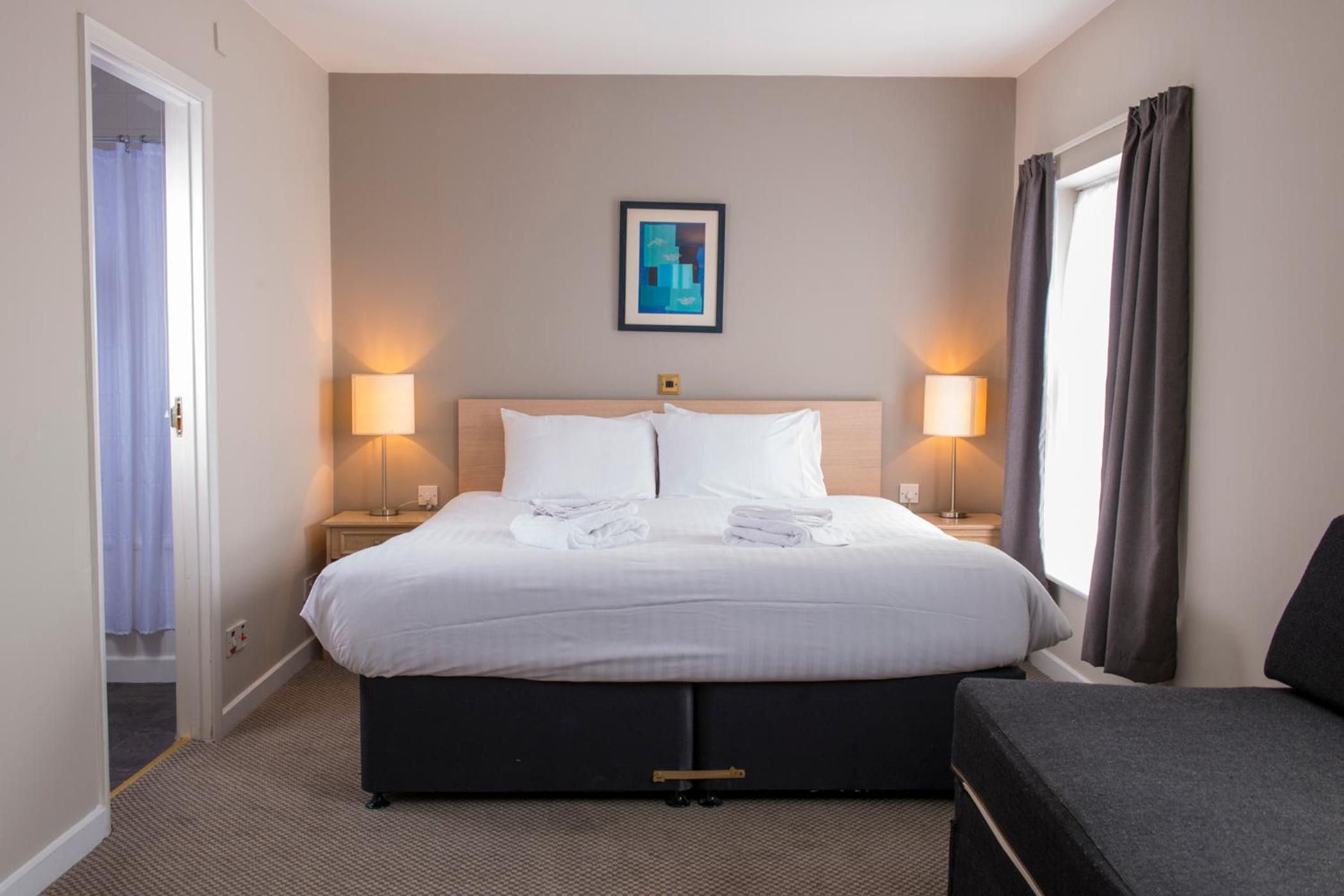 Habitación de hotel con cama, dos lámparas de noche, un cuadro en la pared y una puerta a la izquierda.