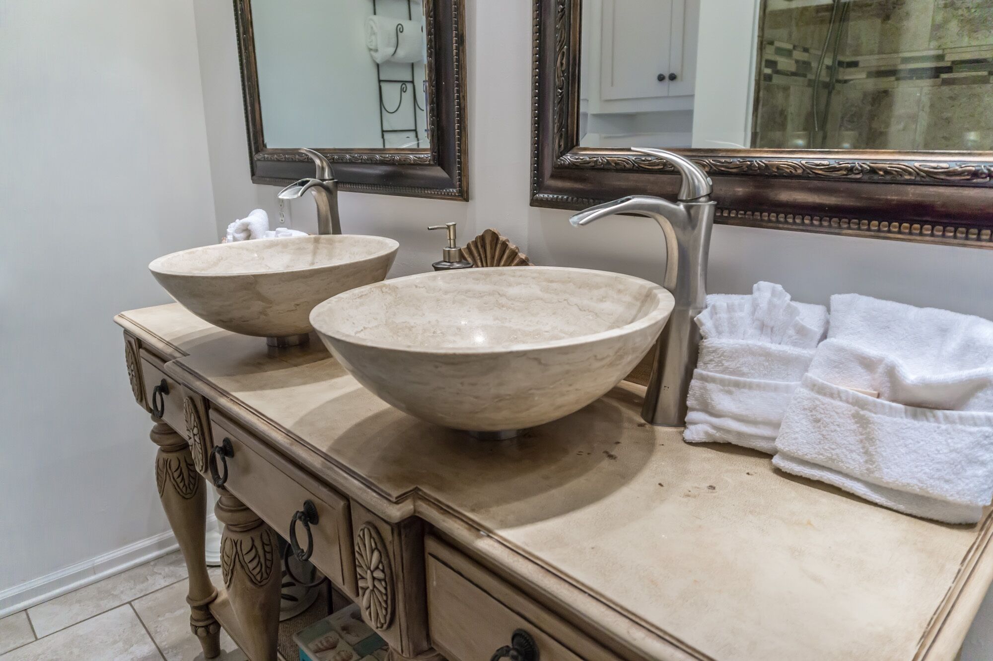 A bathroom with two large, beige, bowl-shaped sinks, a vanity with ornate carvings, and a mirror with an ornate frame.