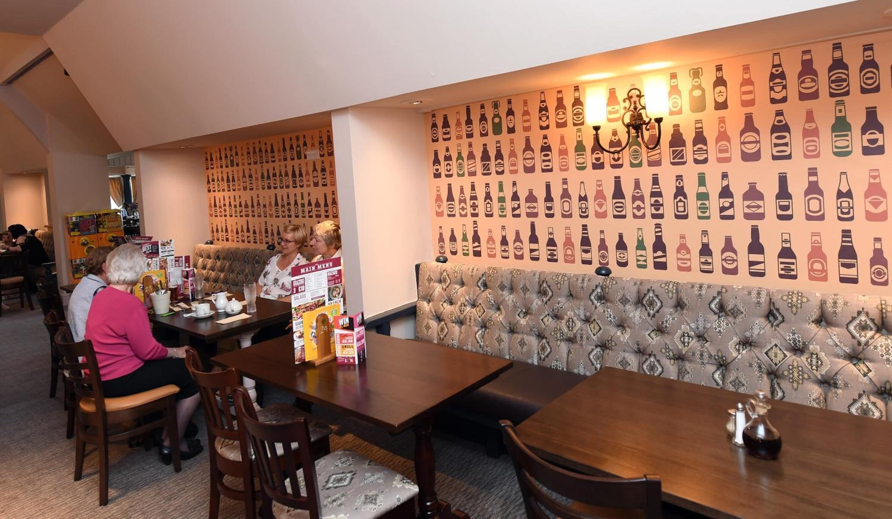 Interior de un restaurante con una pared de iconos de botellas de bebidas, un cabecero capitonado y mesas y sillas de madera.
