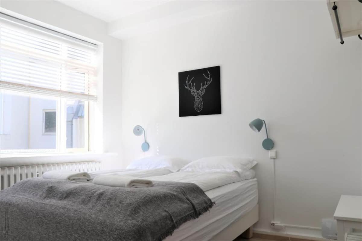 Ein Schlafzimmer mit einem Bett, einem Fenster mit Jalousien, einem Wandbild eines Reh Kopfes und zwei Wandleuchten.