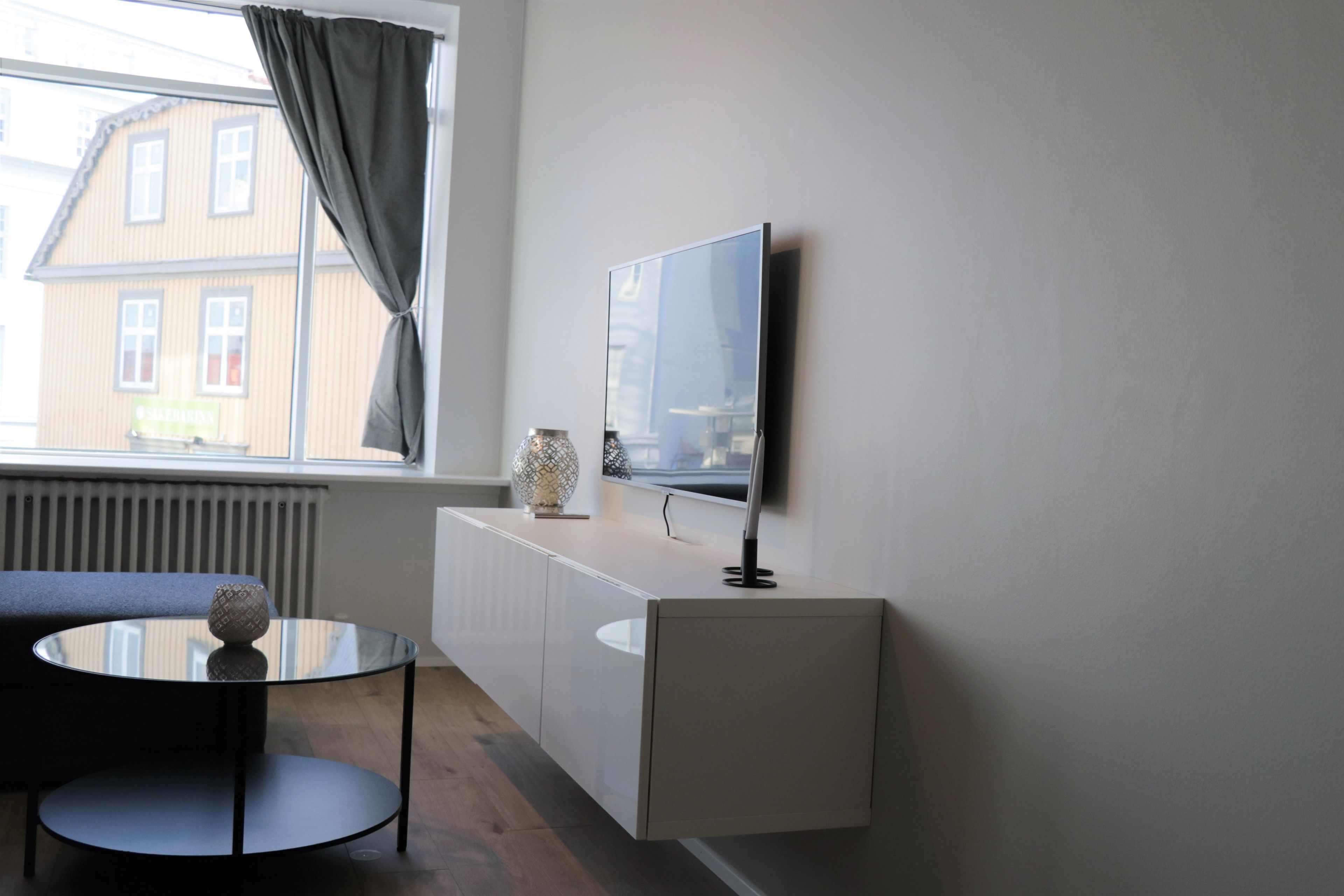 Una habitación moderna con una mesa de vidrio, un sofá, un televisor montado en la pared y una ventana con vistas a edificios.