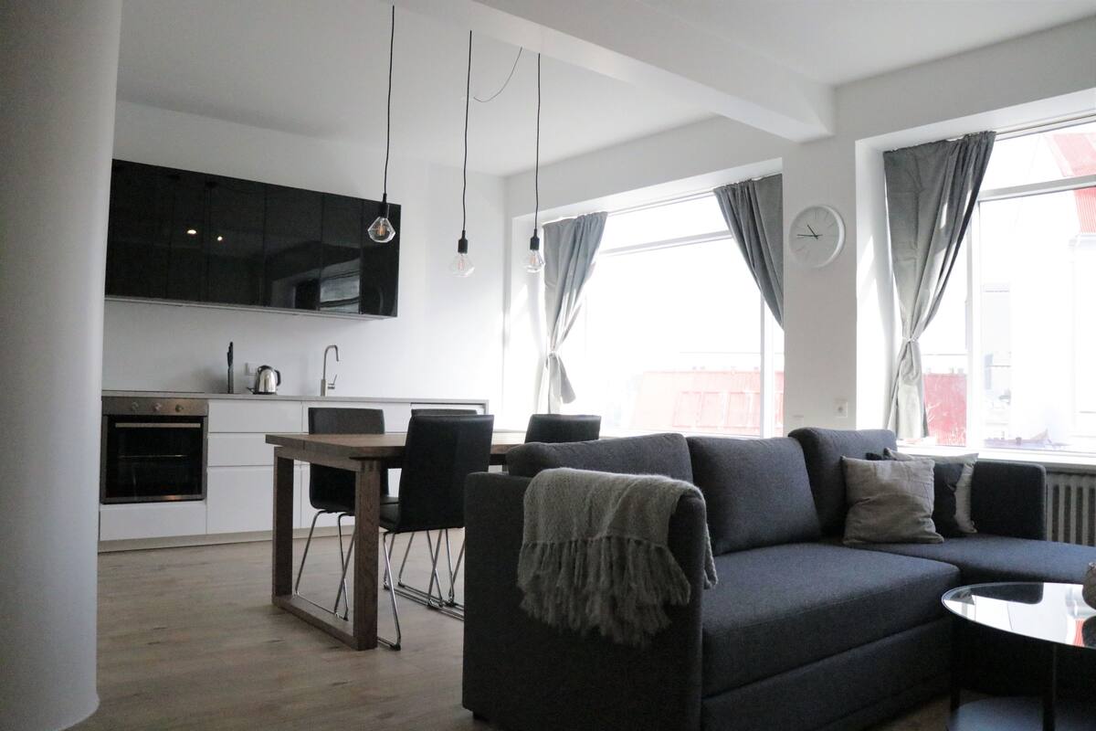Ein modernes Wohnzimmer mit einer dunklen Couch, einem kleinen runden Tisch und einem Essbereich mit einem Holztisch und Stühlen. Im Hintergrund ist eine Küche mit einem schwarzen Schrank und einem Herd erkennbar.