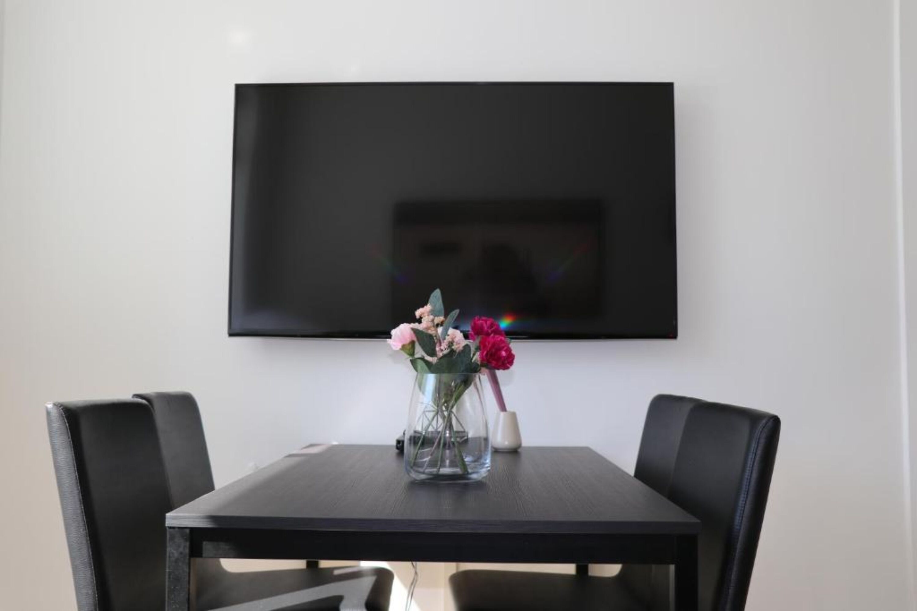 Un coin repas avec une table et des chaises, un vase rempli de fleurs et un téléviseur à écran plat fixé au mur.