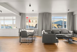 A modern living room with a sofa, chairs, a coffee table, and a dining area with a table and chairs. Large windows offer views of buildings outside.