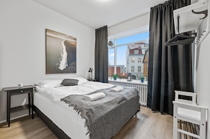 A modern bedroom with a large bed, a nightstand, a window with curtains, and a view of buildings outside.