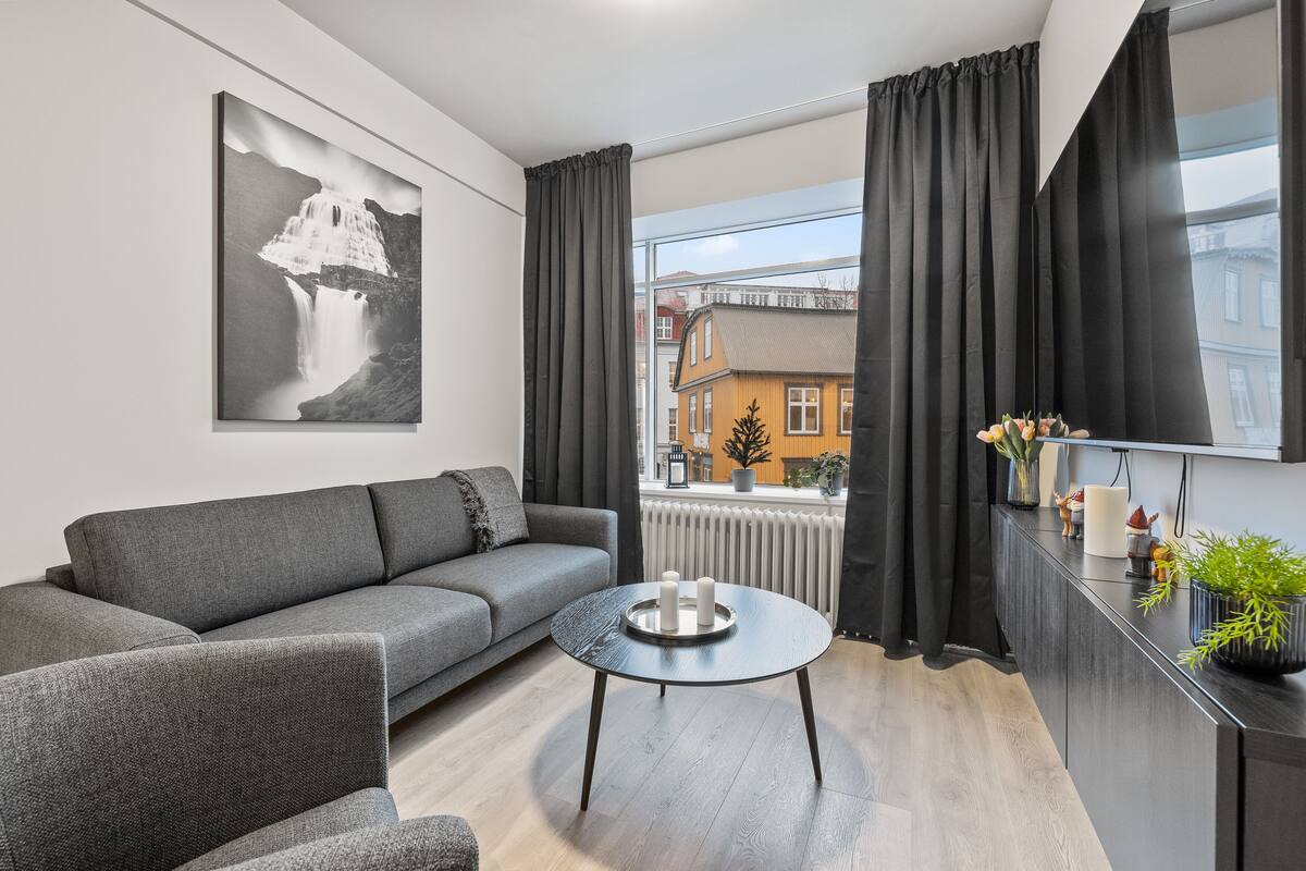 Ein modernes Wohnzimmer mit einem grauen Sofa, einem runden Couchtisch mit Kerzen und einem großen Fenster mit Vorhängen.