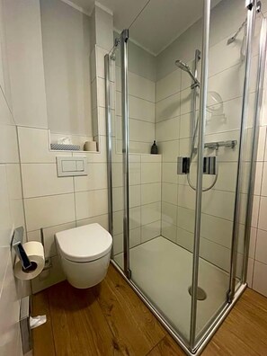 A modern bathroom with a glass-enclosed shower, a toilet, and a small shelf with toiletries.