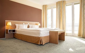 A hotel room with a large bed, bedside lamps, a wooden bench, and a view of the outdoors through a large window.