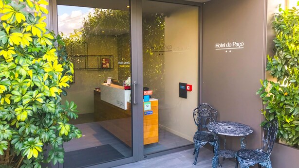 Zona de recepción de un hotel con puerta de vidrio, mostrador de recepción y un pequeño espacio al aire libre con dos sillas y una mesa.