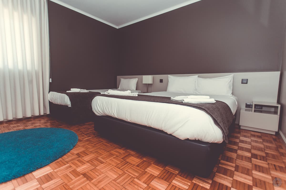 Quarto de hotel moderno com duas camas, tapete azul, piso de madeira e mesa de cabeceira branca.
