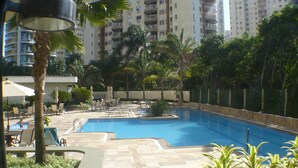 A swimming pool area with lounge chairs and umbrellas, surrounded by palm trees and a high-rise building in the background. - Barra Dolce Vita Residence Service (Rio de Janeiro)