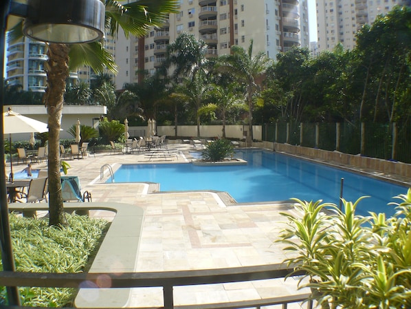 Zona de piscina con sillones de descanso y sombrillas, rodeada de palmeras y un rascacielos al fondo.