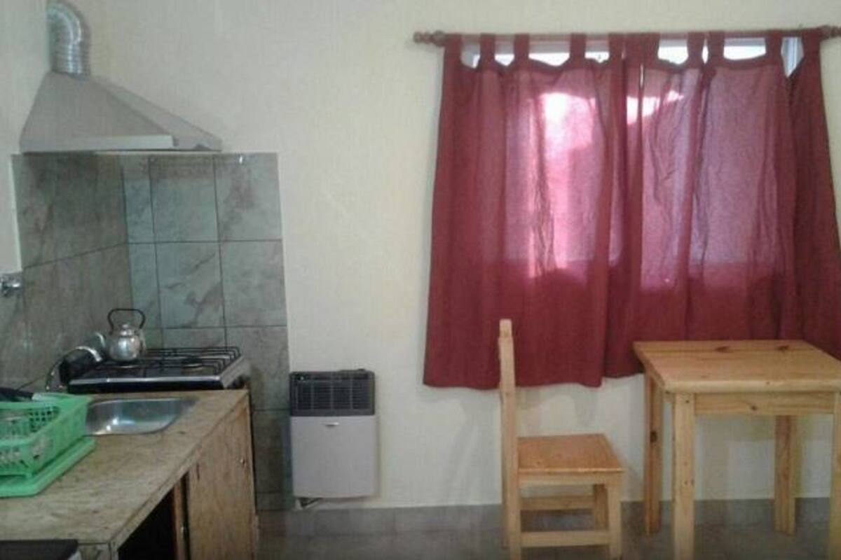 A small kitchen with a gas stove, a kettle, and a microwave. There is a wooden table with two chairs and a red curtain.