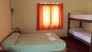 Ein kleines Zimmer mit einem Einzelbett und einem Schlafplatz darüber. Rechts befindet sich ein Etagenbett aus Holz. Ein Fenster mit orangenen Vorhängen, ein Nachttisch mit Lampe und eine Uhr an der Wand sind ebenfalls vorhanden.