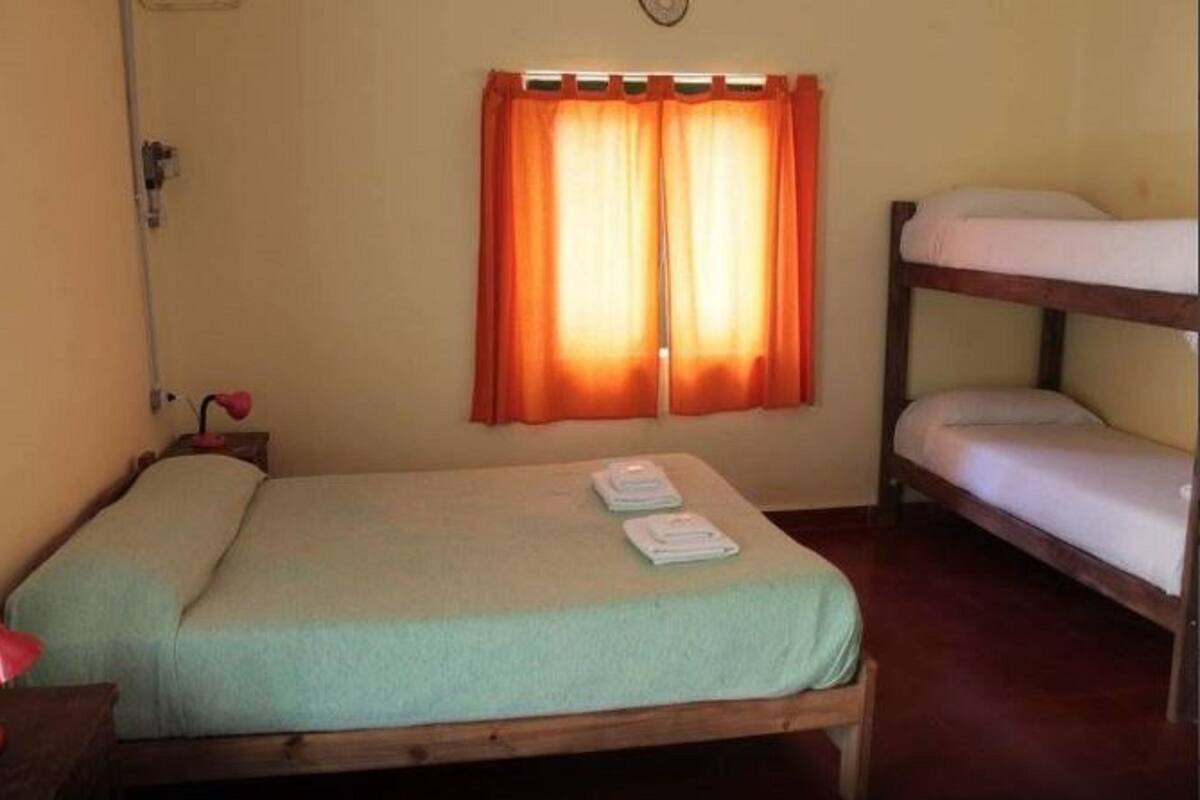 A small, single-bed room with a wooden bunk bed to the right. There is a window with orange curtains, a bedside table with a lamp, and a clock on the wall.