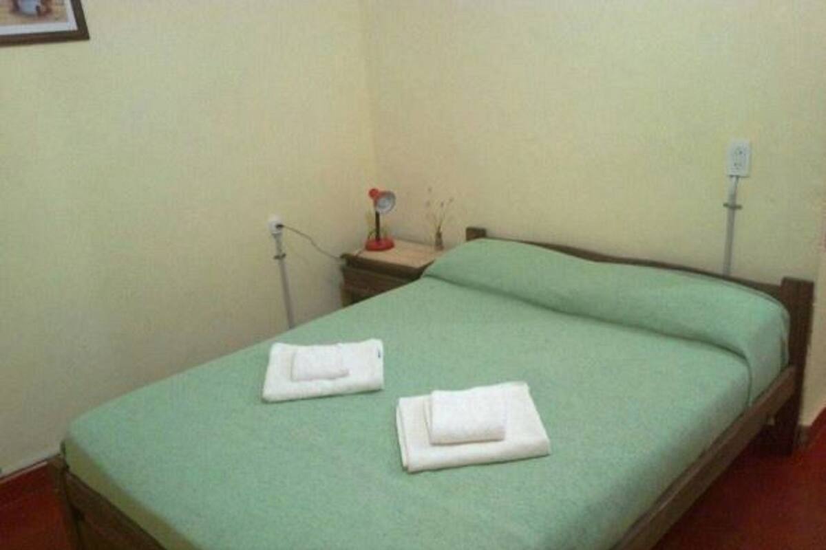 A single-bed room with a green bedspread, two neatly folded white towels, a bedside table with a lamp, and a wall socket.