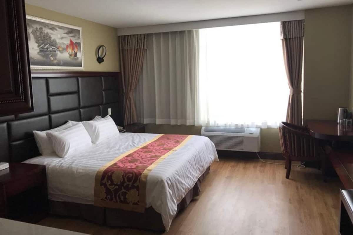 Habitación de hotel con una cama grande, un escritorio y una silla. Se observa una ventana con cortinas, un equipo de aire acondicionado y un cuadro en la pared.