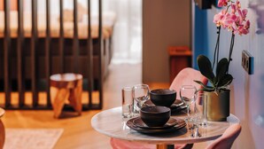 Una mesa de comedor dispuesta con fuentes, vasos y cubiertos, una orquídea en maceta y un taburete pequeño en una habitación con una cama y cortinas al fondo.