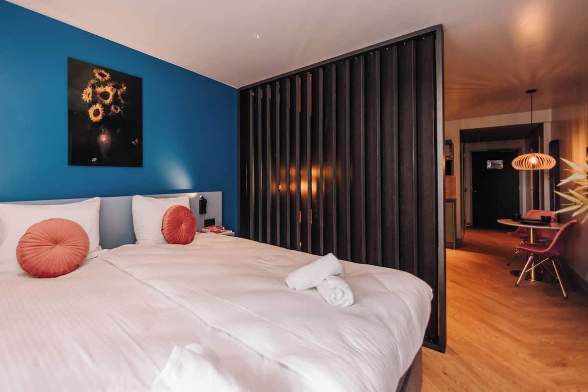 Ein modernes Hotelzimmer mit einem großen Bett, einer blauen Akzentwand, einem Gemälde von Sonnenblumen und einem separaten Essbereich mit Tisch und Stühlen.