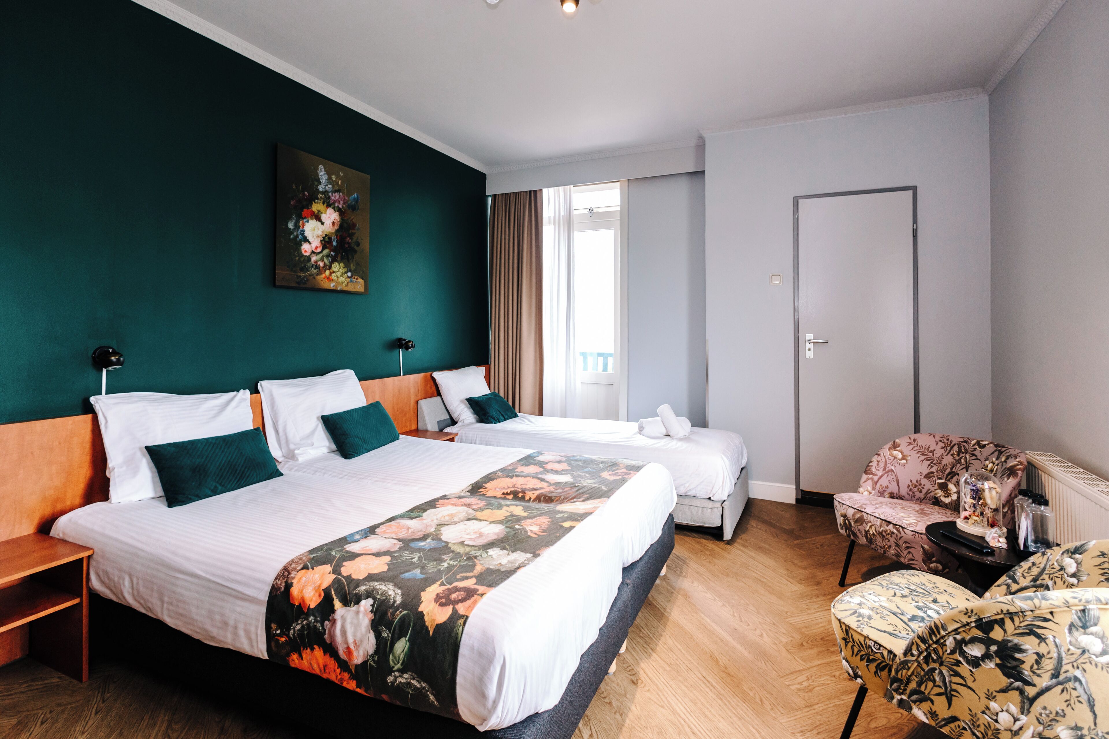 Habitación de hotel con dos camas, colcha estampada, un cuadro en la pared, una silla y vistas al exterior.