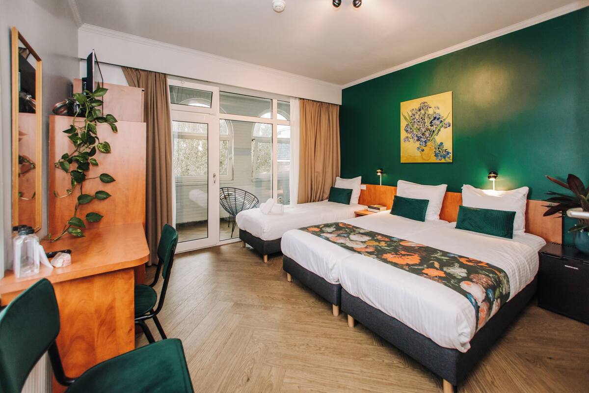 Ein Hotelzimmer mit zwei Betten, einem Schreibtisch und einem Sessel. Ein großes Fenster mit Vorhängen, ein Bild an der Wand und eine Pflanze auf dem Schreibtisch.