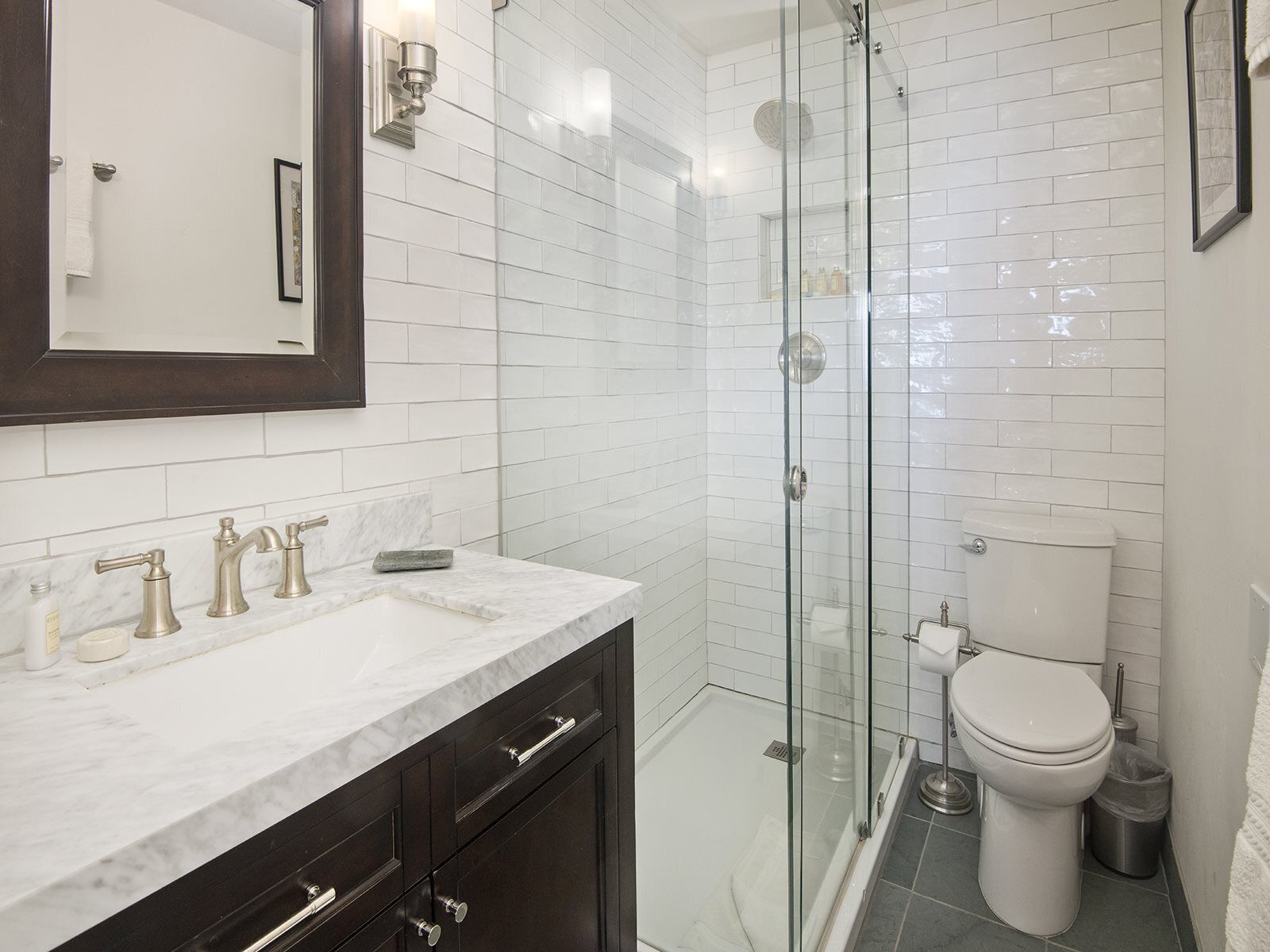 A modern bathroom with a glass-enclosed shower, a white toilet, and a marble sink with dark cabinetry.