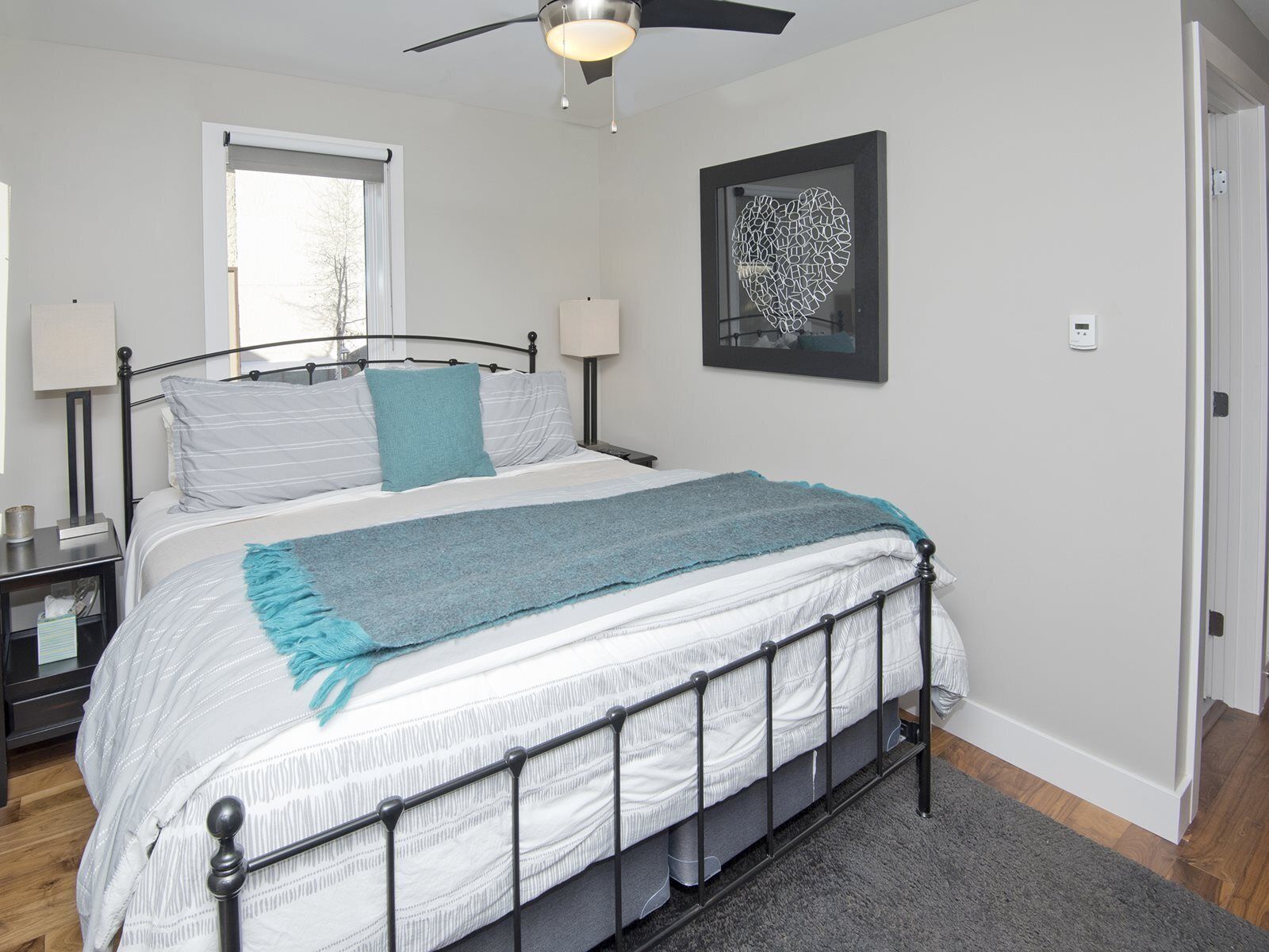 A neatly arranged bedroom with a bed, bedside tables, a ceiling fan, a window, and a wall-mounted artwork.