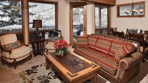 A cozy living room with a sofa, armchair, and coffee table, large windows with blinds, and a chandelier. - Pines Townhome (Avon)