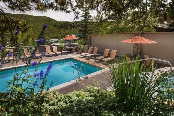 A pool area with lounge chairs and umbrellas, surrounded by greenery and a view of hills.