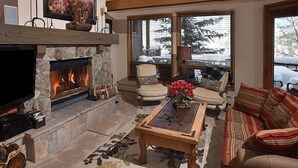 A cozy living room with a stone fireplace, comfortable seating, and a large window offering a view of the outdoors. - Pines Townhome (Avon)