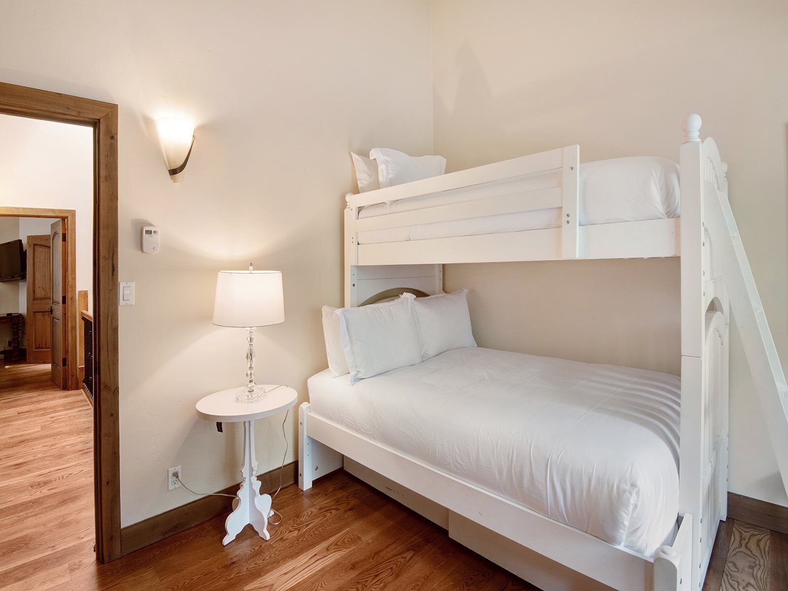 A bunk bed room with a bedside table and lamp, wooden flooring, and a door leading to another room.