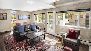 Ein großzügiges Wohnzimmer mit einer Ledersofa, einem Couchtisch und einem roten Teppich mit Muster. Große Fenster bieten Blick auf eine Wohngegend.