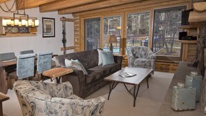 Ein gemütliches Wohnzimmer mit einer Couch, Sesseln und einem Couchtisch, Holzbalken, einem Chandelier und großen Fenstern mit Blick auf Bäume.