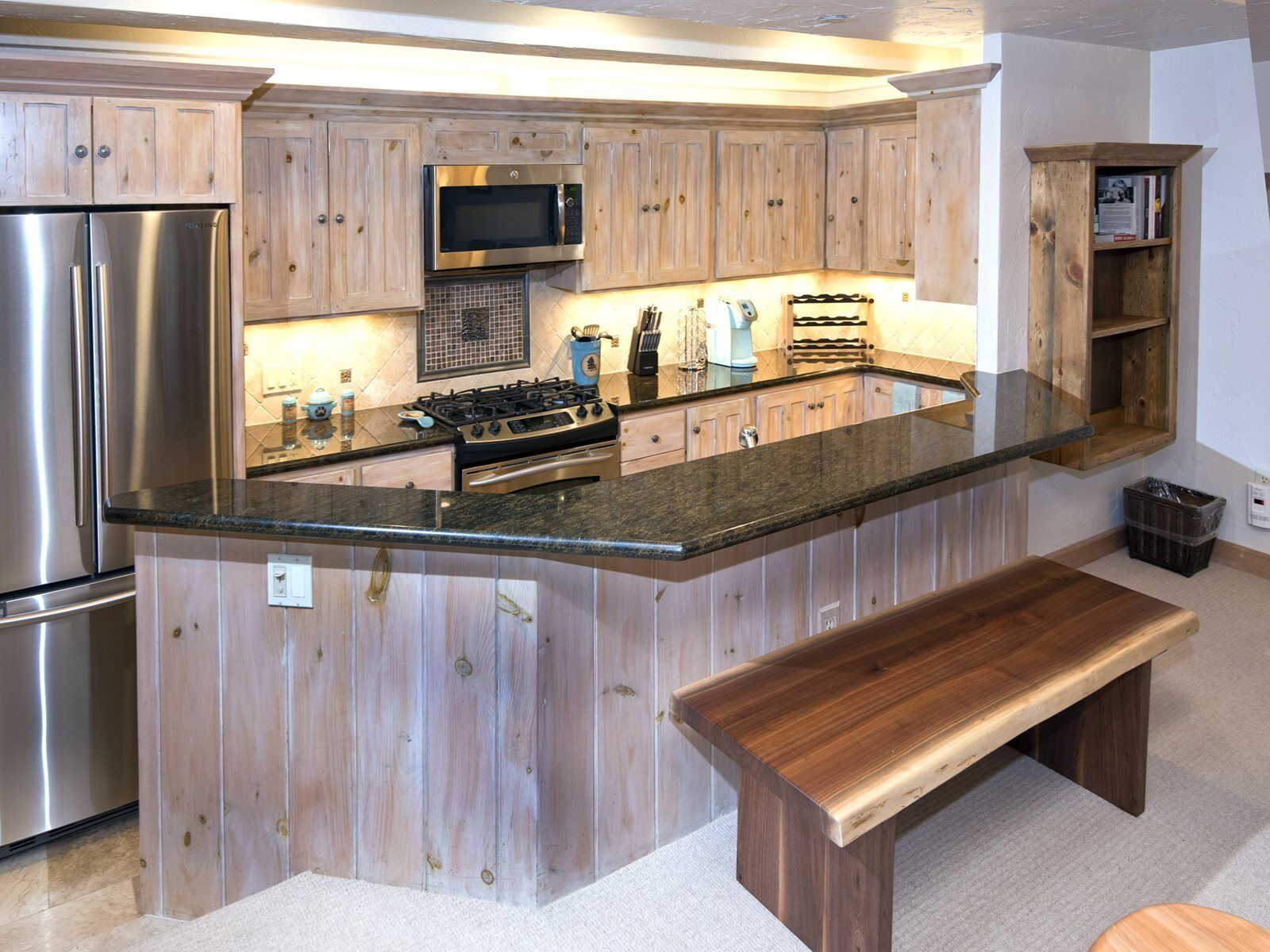 Une cuisine dotée de meubles en bois, d’appareils électroménagers en acier inoxydable, d’un plan de travail en granit et d’un banc en bois.