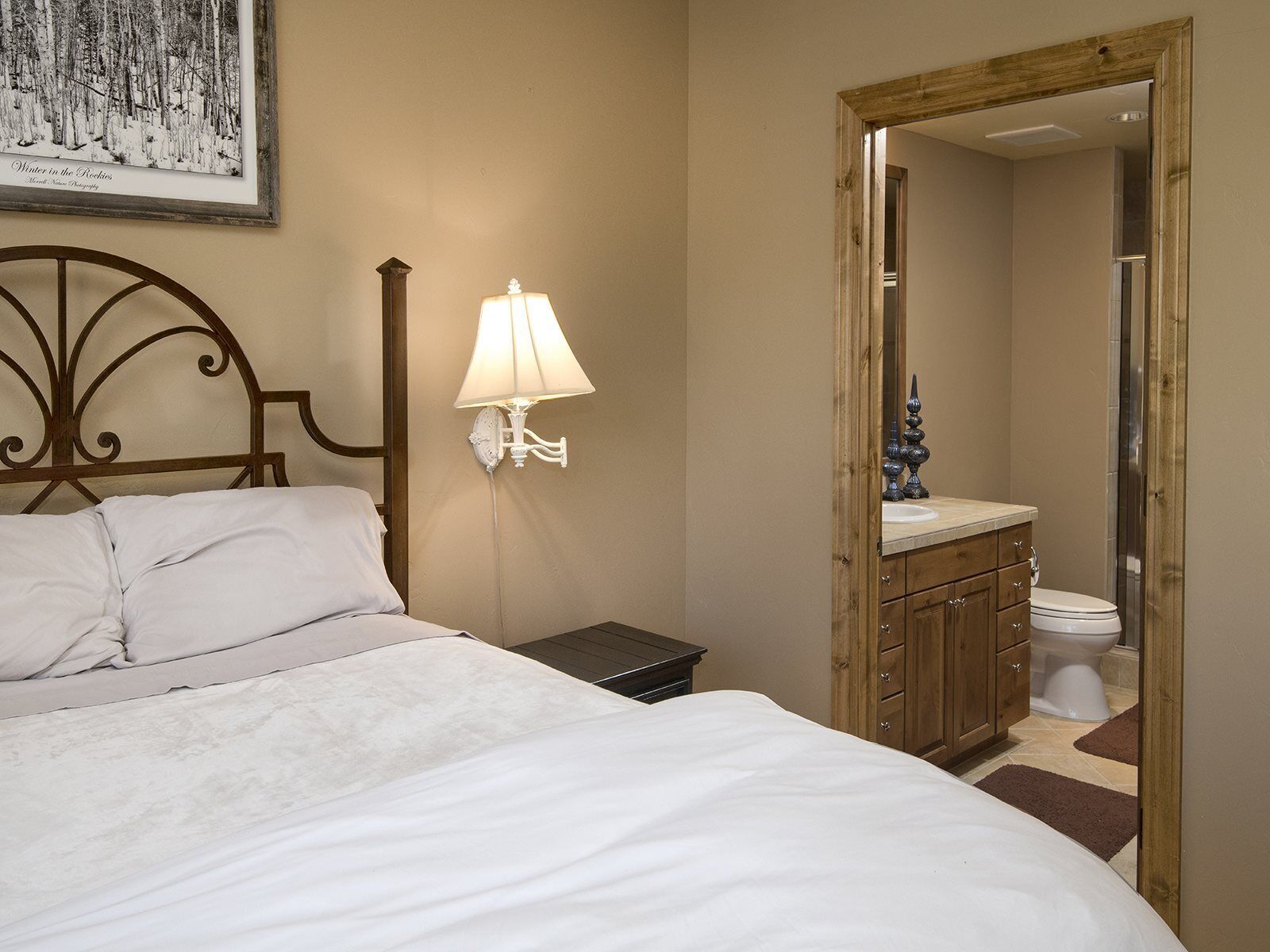 A bedroom with a bed, a nightstand, a wall-mounted lamp, a wooden headboard, a framed picture, and a bathroom visible through an open door.