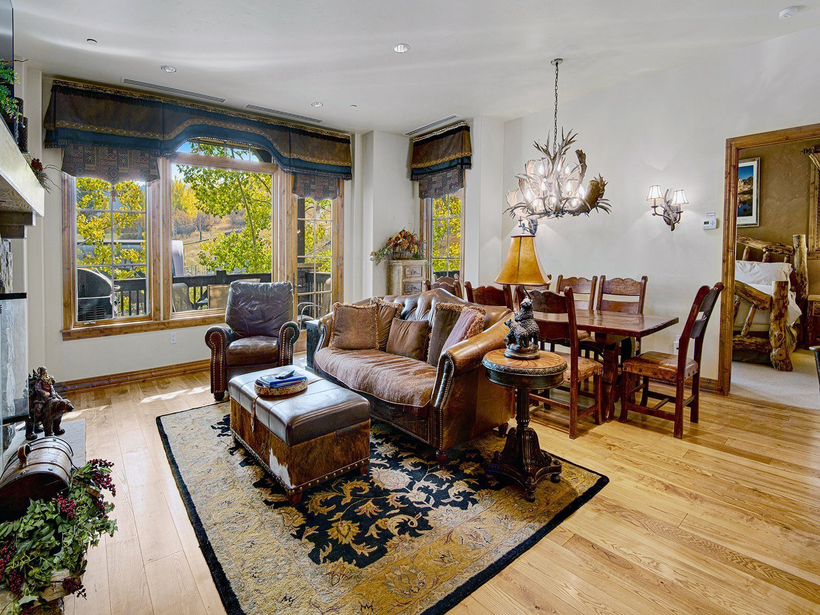 A spacious living room with a leather sofa, a dining table with chairs, and a chandelier. Large windows provide natural light and views of greenery outside.