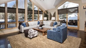 A spacious living room with a large sectional sofa, a blue armchair, and a coffee table. There are large windows offering a view of snow-covered trees. - Glacier Court 1854 A (Vail)