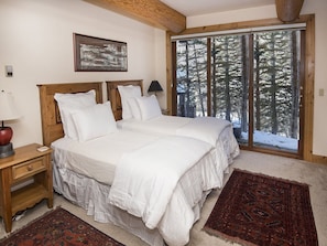 A bedroom with a large bed, wooden headboard, white bedding, a nightstand with a lamp, a red decorative item, a patterned rug, and a window with a view of trees.