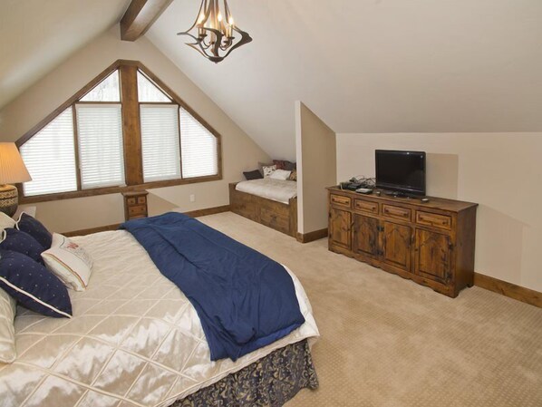 A bedroom with a sloped ceiling, a large bed, a dresser, a television, and a window with blinds.
