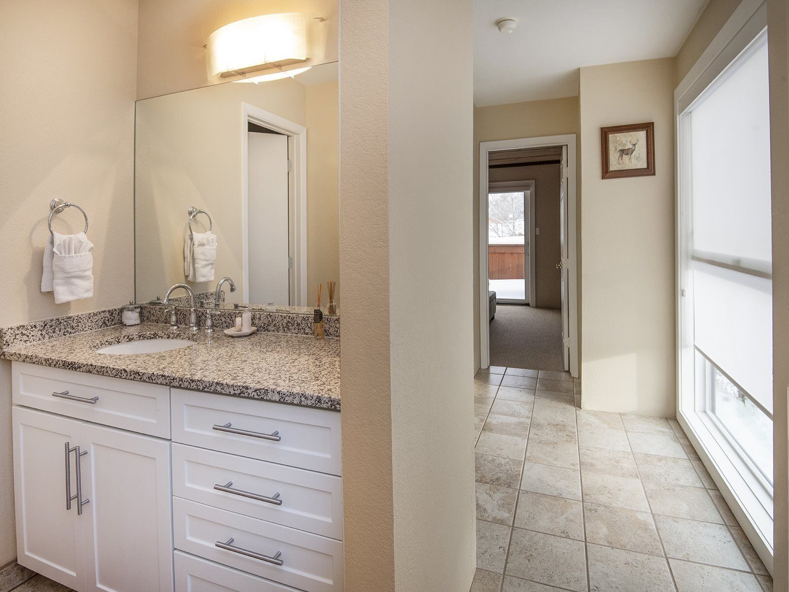A bathroom with a granite countertop, white cabinets, a large mirror, and a framed picture on the wall.