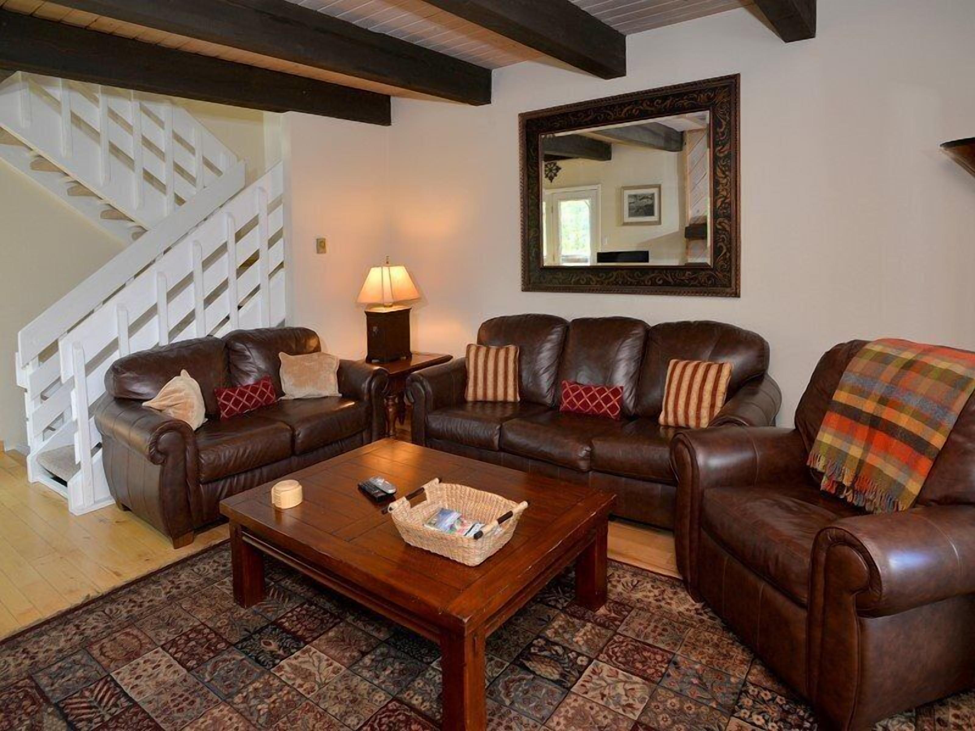 A living room with a brown leather sofa set, a wooden coffee table, and a patterned rug. A white staircase is visible in the background.