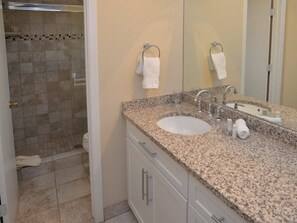 Un baño con lavamanos de encimera de granito, un espejo grande y una zona de ducha con paredes de azulejos.